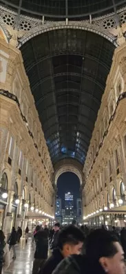 A low angle photo looking up at the high, arched glass ceiling and ornate facades of an architectural gallery in Milan at night.