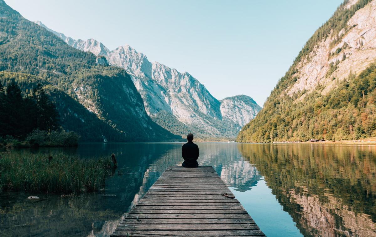 person sitting down at edge of bridge in front of a lake surrounded by mountains