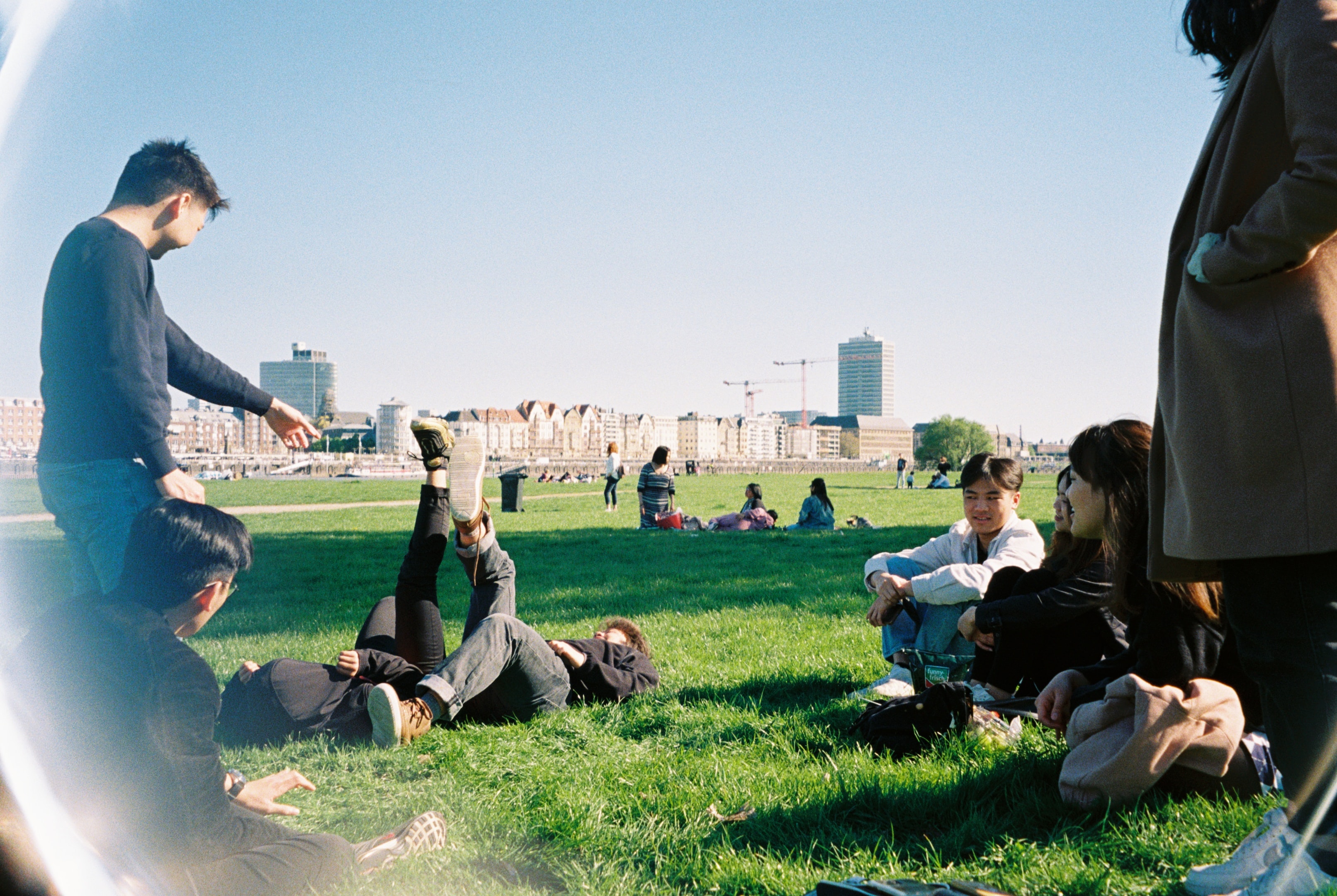 A group of young people hanging out, sitting on the grass in the park with a city behind them.