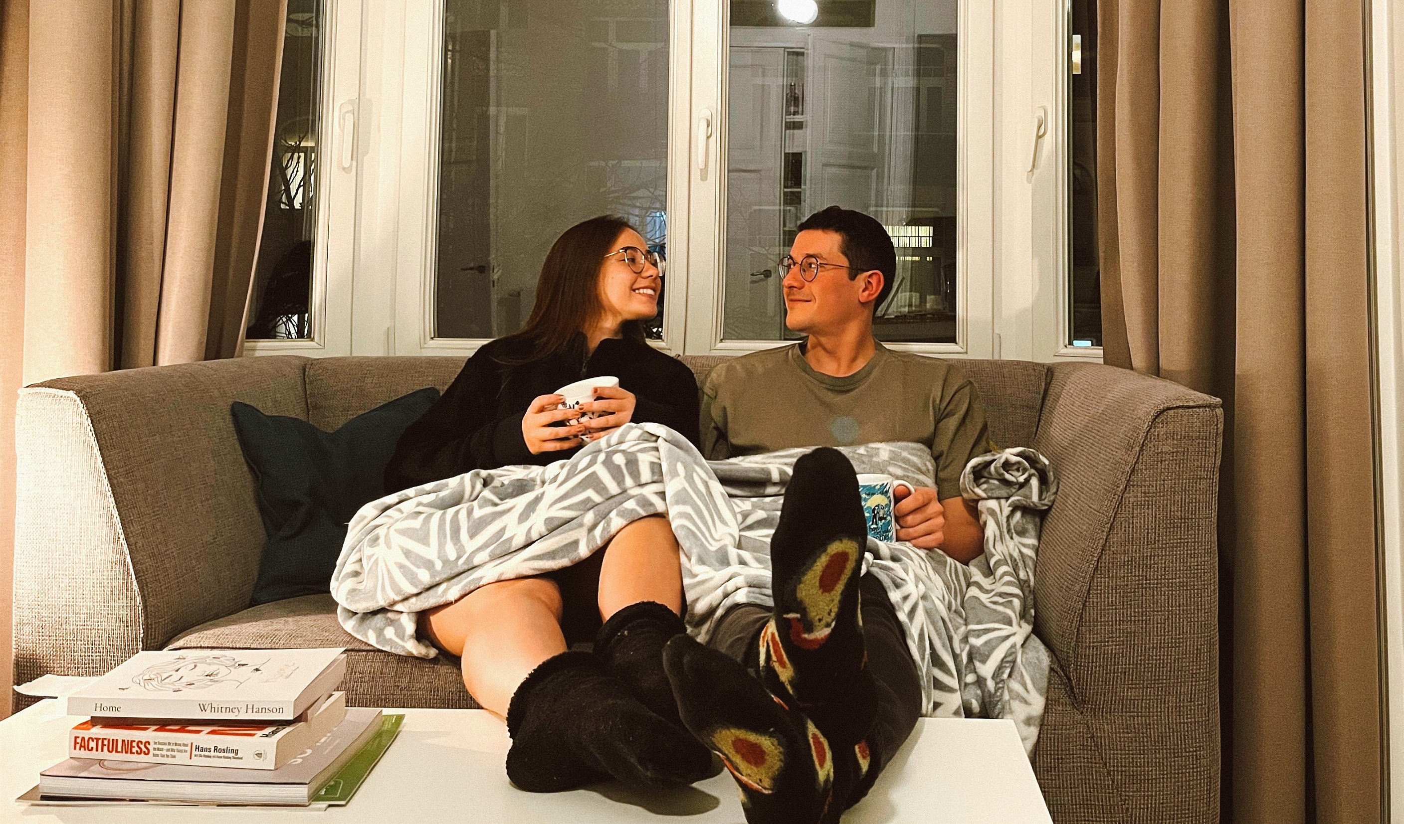 Lala and Nic sitting together on a couch. They are wrapped up in a gray and white blanket, with mugs, looking at each other. Their feet are propped up on a table, with some books on the side.