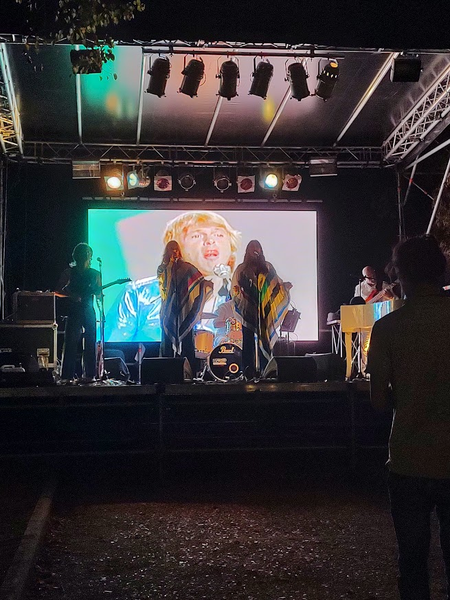 A stage with four people dressed as musicians from the band Abba. In the background a picture of one of the musicians.