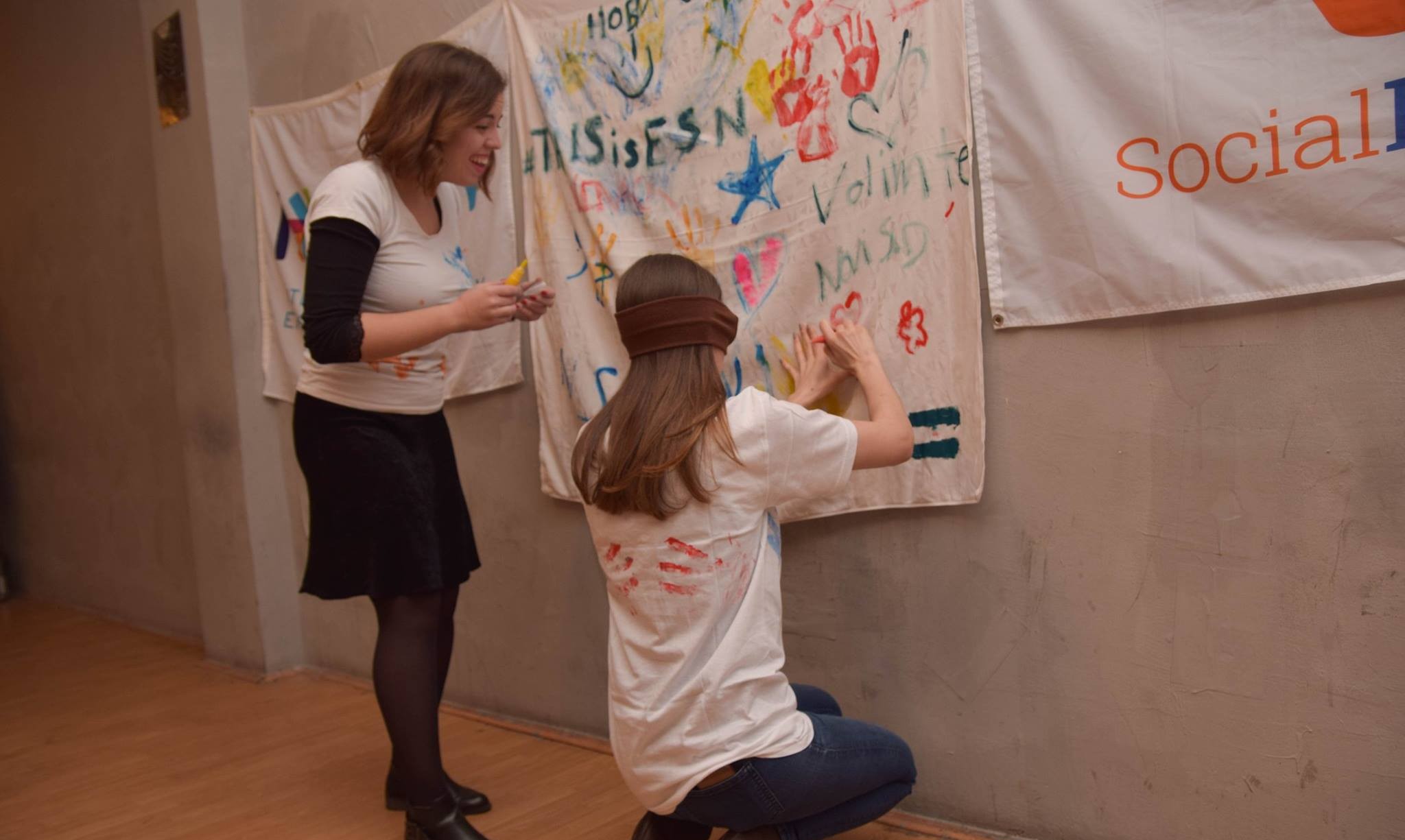 Two people, one blindfolded and painting on a white fabric on the wall