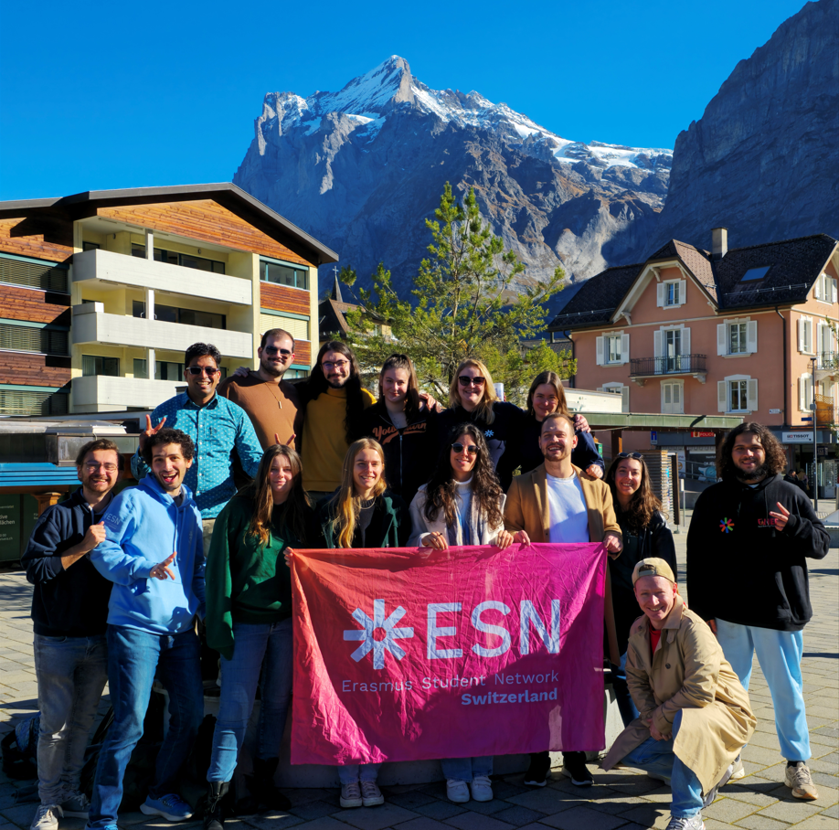 Image of 15 Swiss national level members smiling at the camera in front of a mountain and holding the ESN Switzerland flag.