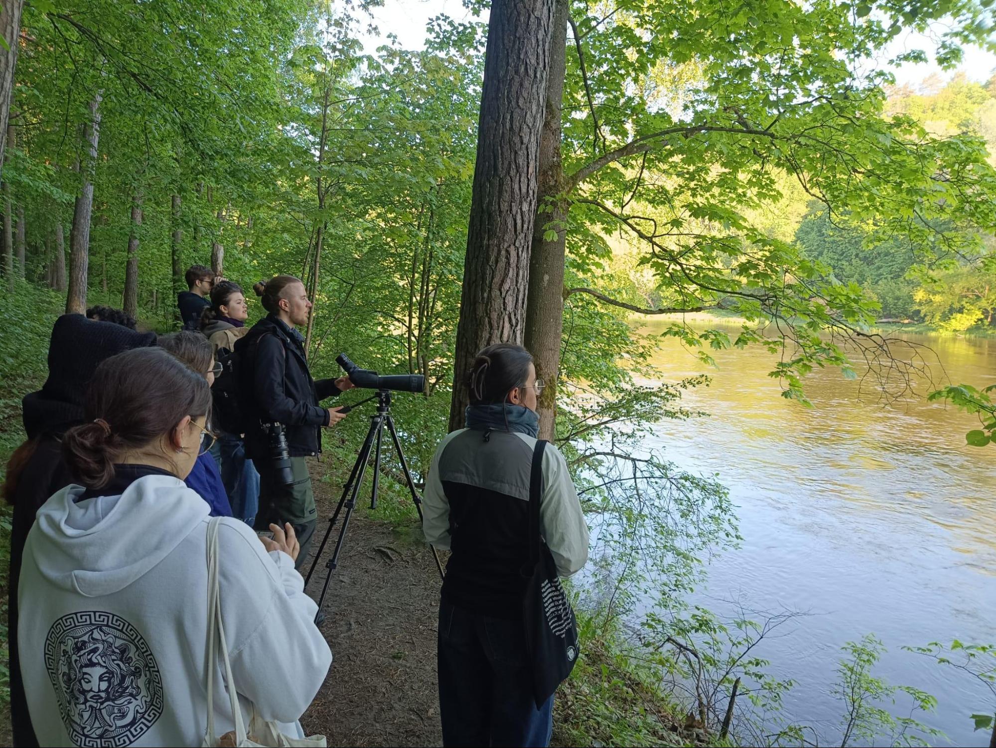 The image shows some people in front of a river in the forest with a telescope seeingbirds