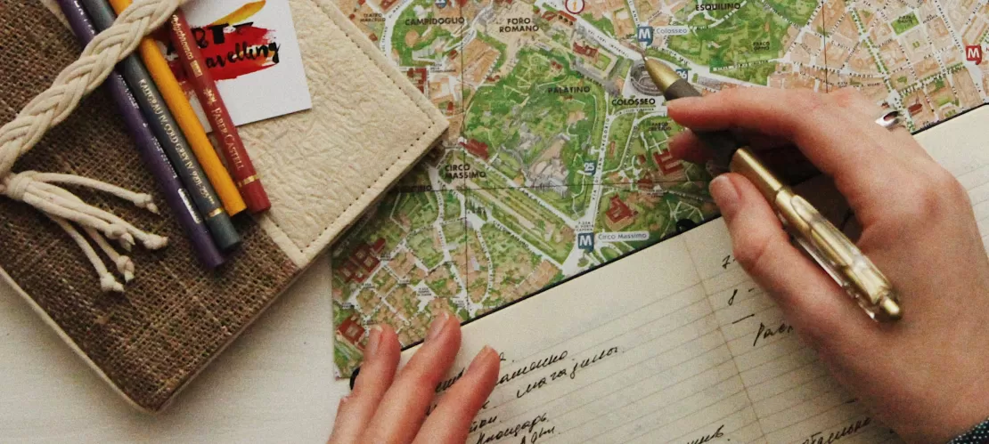 a girl writing in a journal with an open mape of Rome and a book with pencils next to it