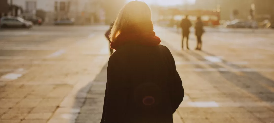 A woman with her back turned towards the camera, looking into the distance. She is standing on a city square with a sunset in the distance.