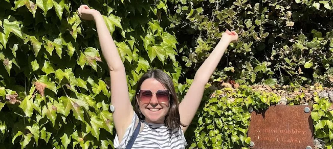 A young woman standing wearing a white and blue-striped t-shirt, with visible insulin pump, standing with her hands up in front of a plant-covered wall. Next to her a brown plaque with unreadable text.