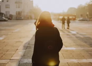 A woman with her back turned towards the camera, looking into the distance. She is standing on a city square with a sunset in the distance.