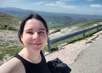 A dark-haired smiling girl standing on a path with a view of mountains in the background.