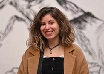 A girl with brown hair and in a light brown coat smiling at the camera.