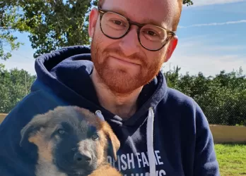 A ginger person with glasses and short bear holding a puppy of belgian malinois.