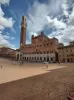 A tall orange-brown building (Palazzo Publico in Siena) with a high tower behind it (Torre del Mangia). In the front there is a square (Piazza del Campo) and few people walking.
